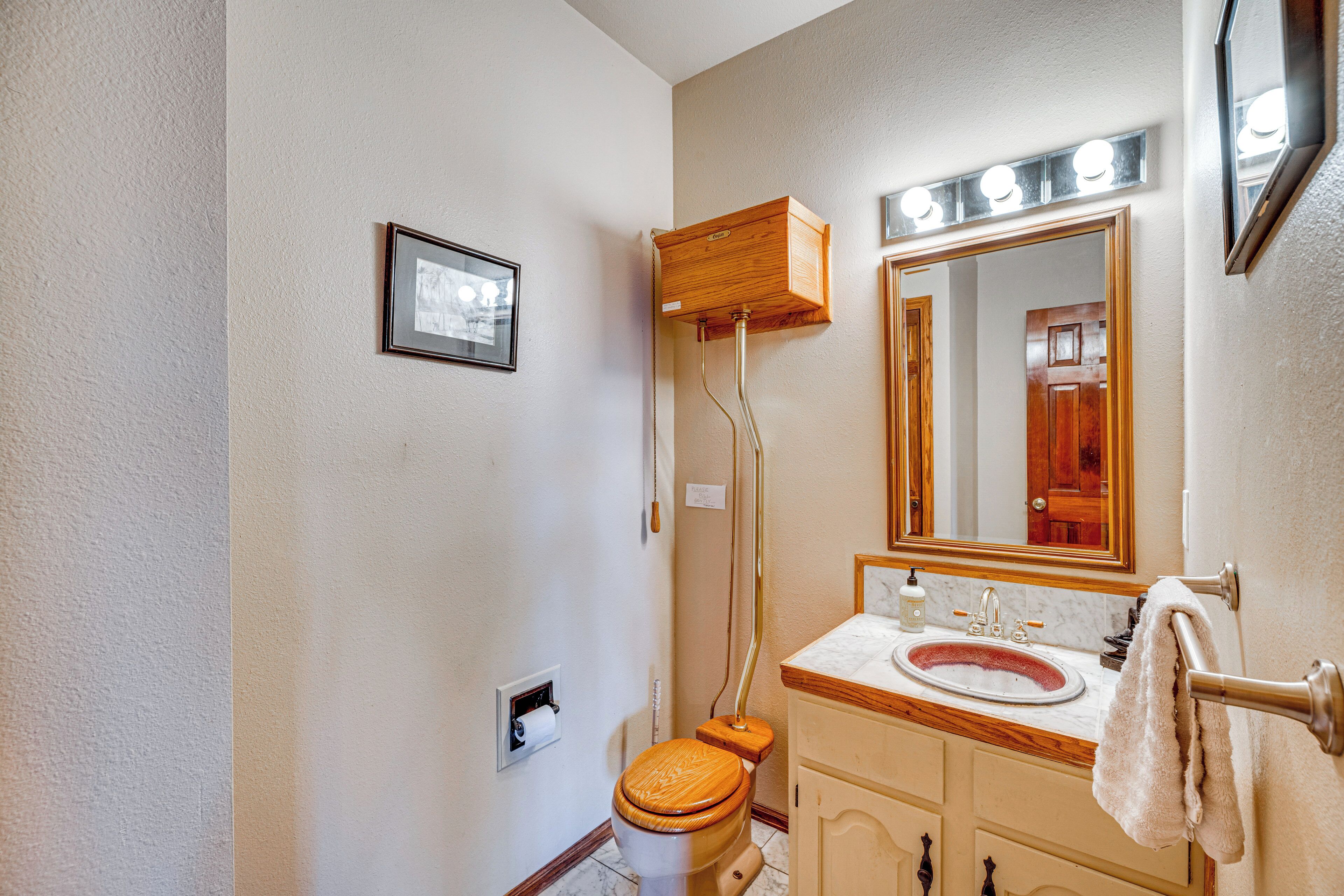 Un baño con inodoro de madera, lavamanos con encimera de mármol, espejo con iluminación y un perchero para toallas.