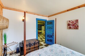Una habitación con una cama, una puerta azul, suelo a cuadros, una silla de mimbre y una lámpara de pared.