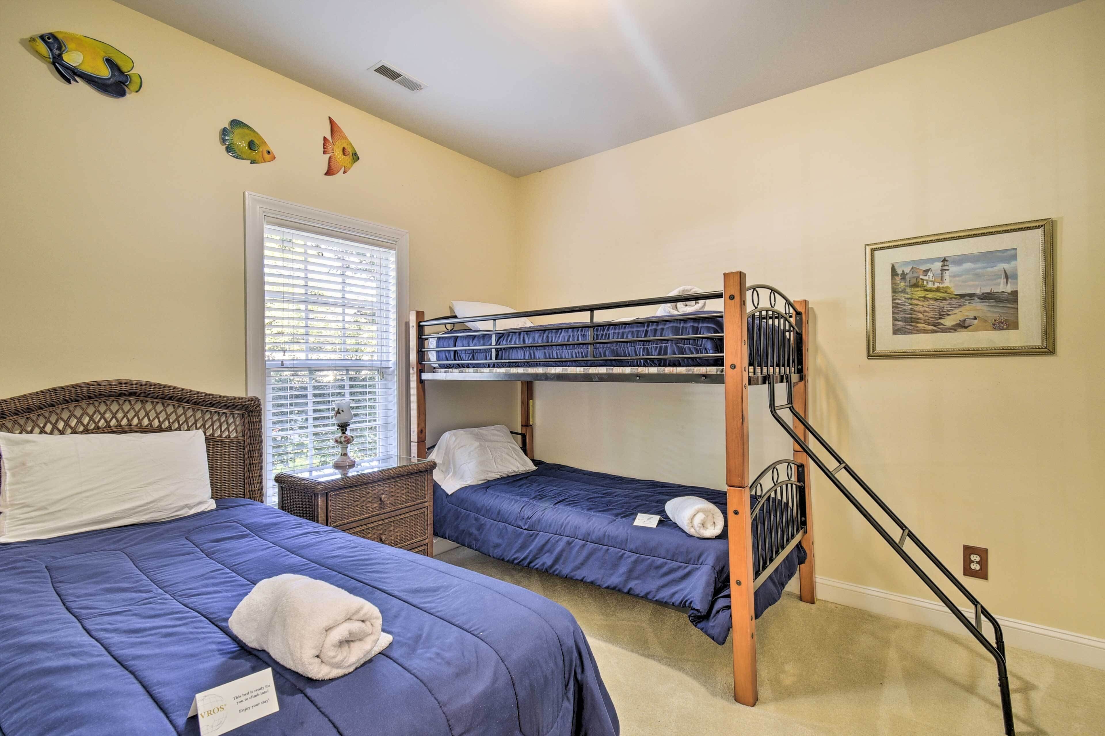 A bedroom with two beds, a bunk bed, a window with blinds, a painting on the wall, and a small table with a lamp.