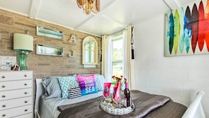 A bedroom with a brick wall, a bed with a dark brown bedspread, a bedside table with a lamp, and a wall-mounted mirror. - Oceanfront White Stone Cottage w/ Private Beach! (White Stone)