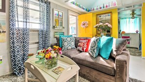 A living room with a brown sofa, teal and white patterned pillows, a glass coffee table with a vase of flowers, and a yellow wall with a mirror and a 'HAPPY' sign. - Oceanfront White Stone Cottage w/ Private Beach! (White Stone)