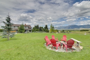 A grassy backyard with a fire pit and red chairs, a house in the background, and mountains in the distance. - Bozeman Home on 11 Acres w/ Mountain Views! (Bozeman)