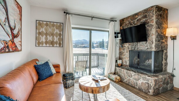 A living room with a stone fireplace, a large window with curtains, a leather sofa, and a flat-screen TV mounted on the wall. - Condo w/ Balcony & Views - Steps to Ski Shuttle! (Fraser)