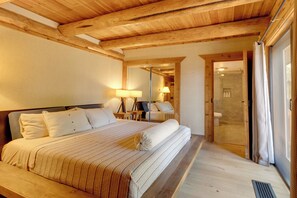 A bedroom with a wooden ceiling, a large bed with white bedding, a bedside table with a lamp, and a bathroom visible through an open door. - Hand-crafted Cabin w/ Whitefish Lake Views! (Whitefish)
