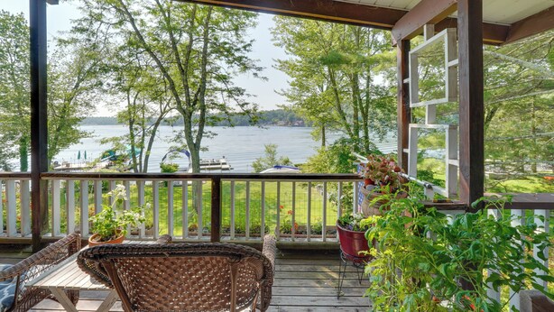 A covered deck with wicker furniture, potted plants, and a view of a lake and trees. - Waterfront White Lake Vacation Rental! (Kauneonga Lake)