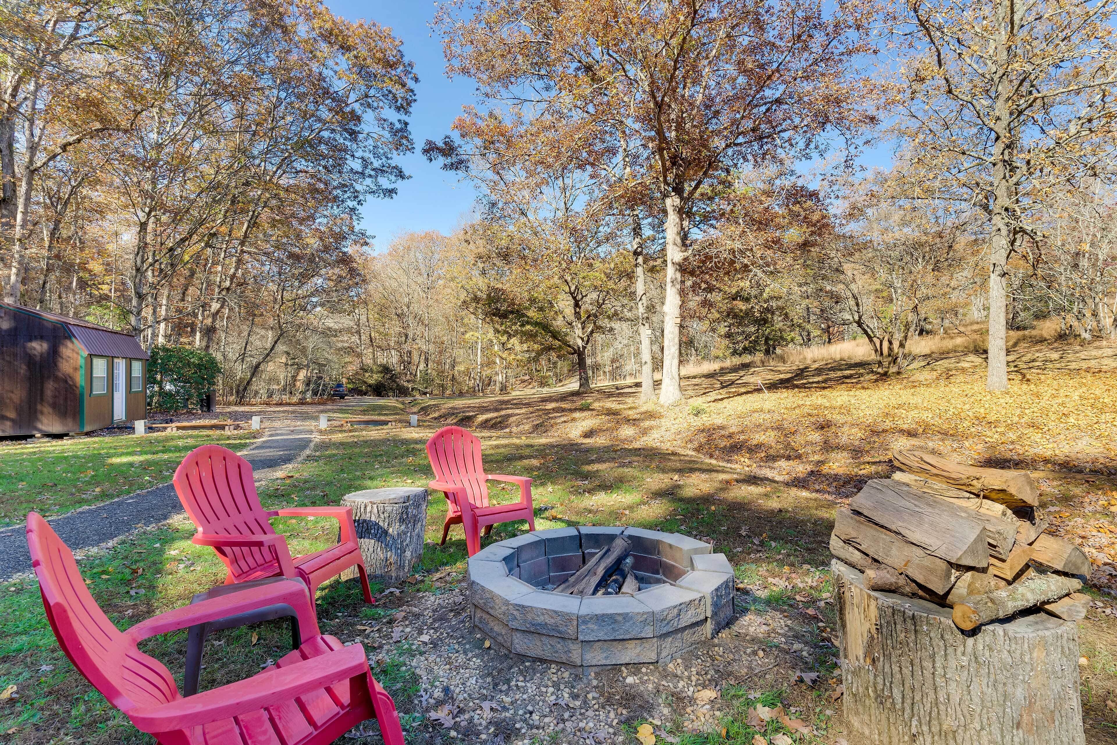 A fire pit with two Adirondack chairs and a stack of firewood in a wooded area.