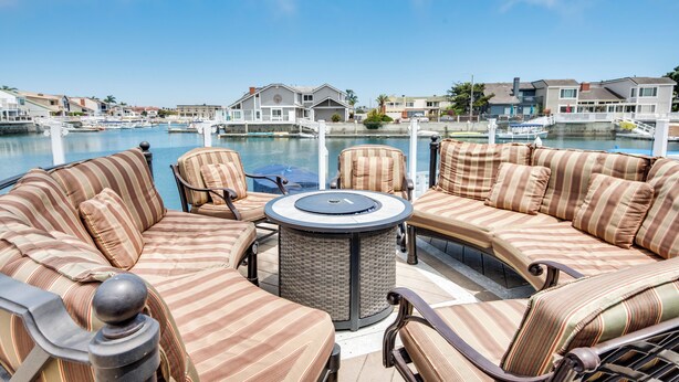 A waterfront patio with striped sofas and a round table, overlooking a marina and residential buildings. - Luxurious Channel Islands Harbor Home w/ Boat Dock (Oxnard)