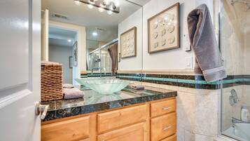 Une salle de bain avec un lavabo en forme de bol en verre, des meubles en bois, une cabine de douche en verre et des œuvres d’art encadrées accrochées au mur.