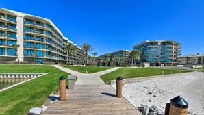 Un camí de fusta cap a un hotel a la platja, amb diversos balcons i palmeres.