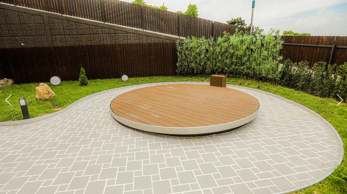 A circular wooden deck with a brick border, surrounded by a grassy area and a wooden fence.