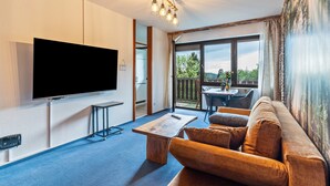 A modern living room with a flat-screen TV, a wooden coffee table, a sofa, and a balcony with a view of trees.