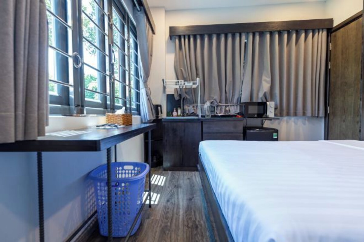 Une chambre d’hôtel avec un lit, un bureau avec un ordinateur, une petite table avec un panier, un panier à linge et une fenêtre avec des rideaux.