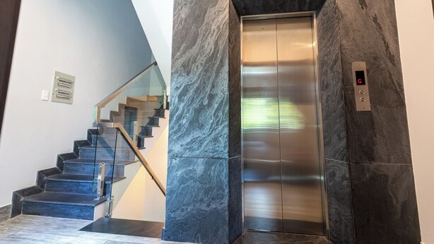 A modern elevator with a metallic finish, a glass staircase with metal railings, and a marble wall.