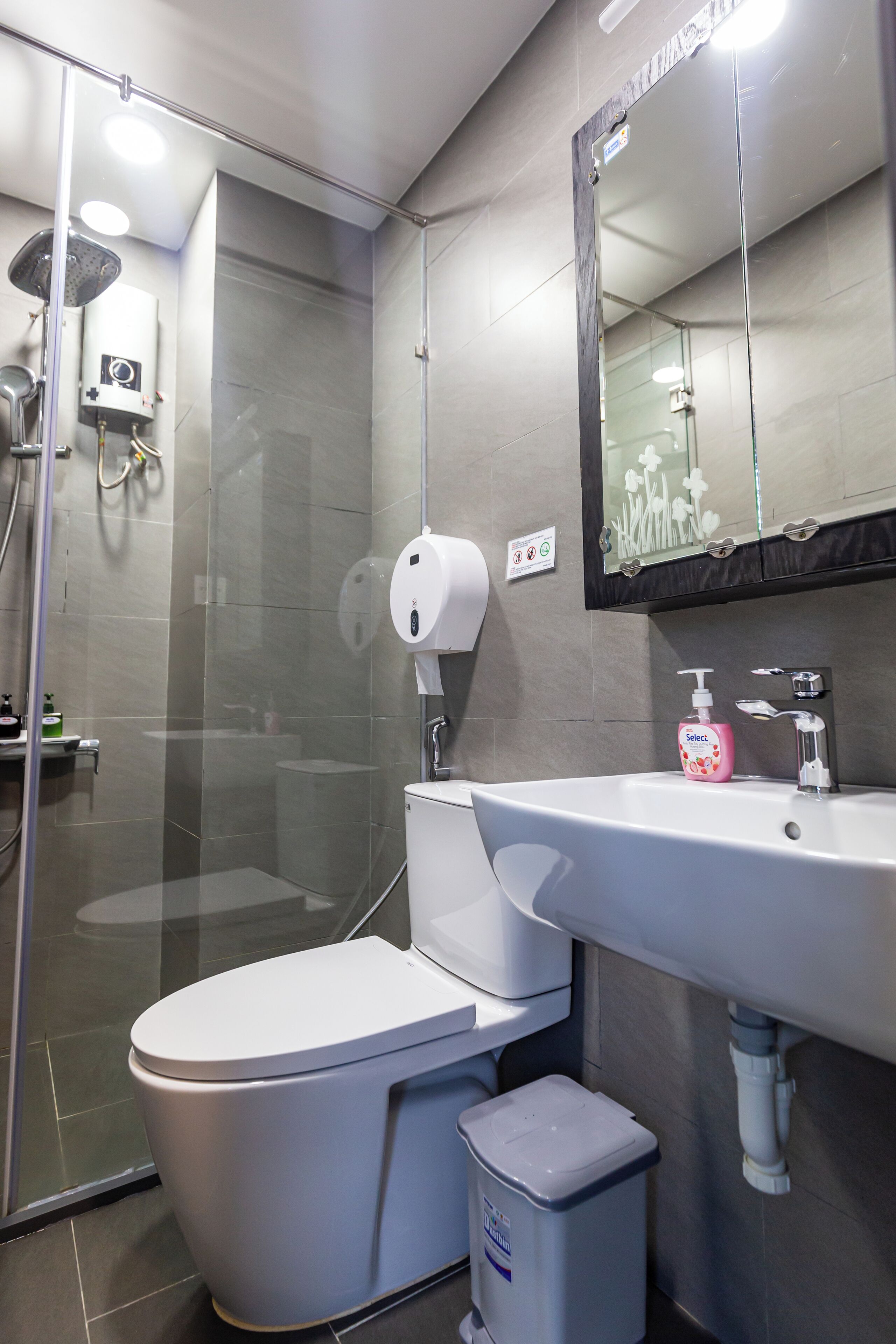 A modern bathroom with a glass-enclosed shower, a white toilet, a sink with a soap dispenser, and a trash bin.