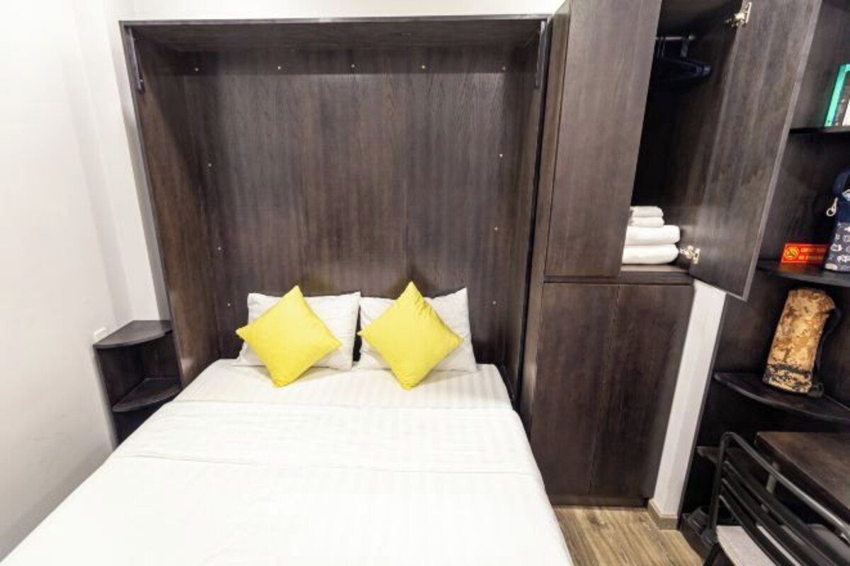 Une chambre à coucher avec un lit, deux oreillers jaunes, une tête de lit en bois foncé, une table de chevet et une étagère avec des serviettes et un objet décoratif.