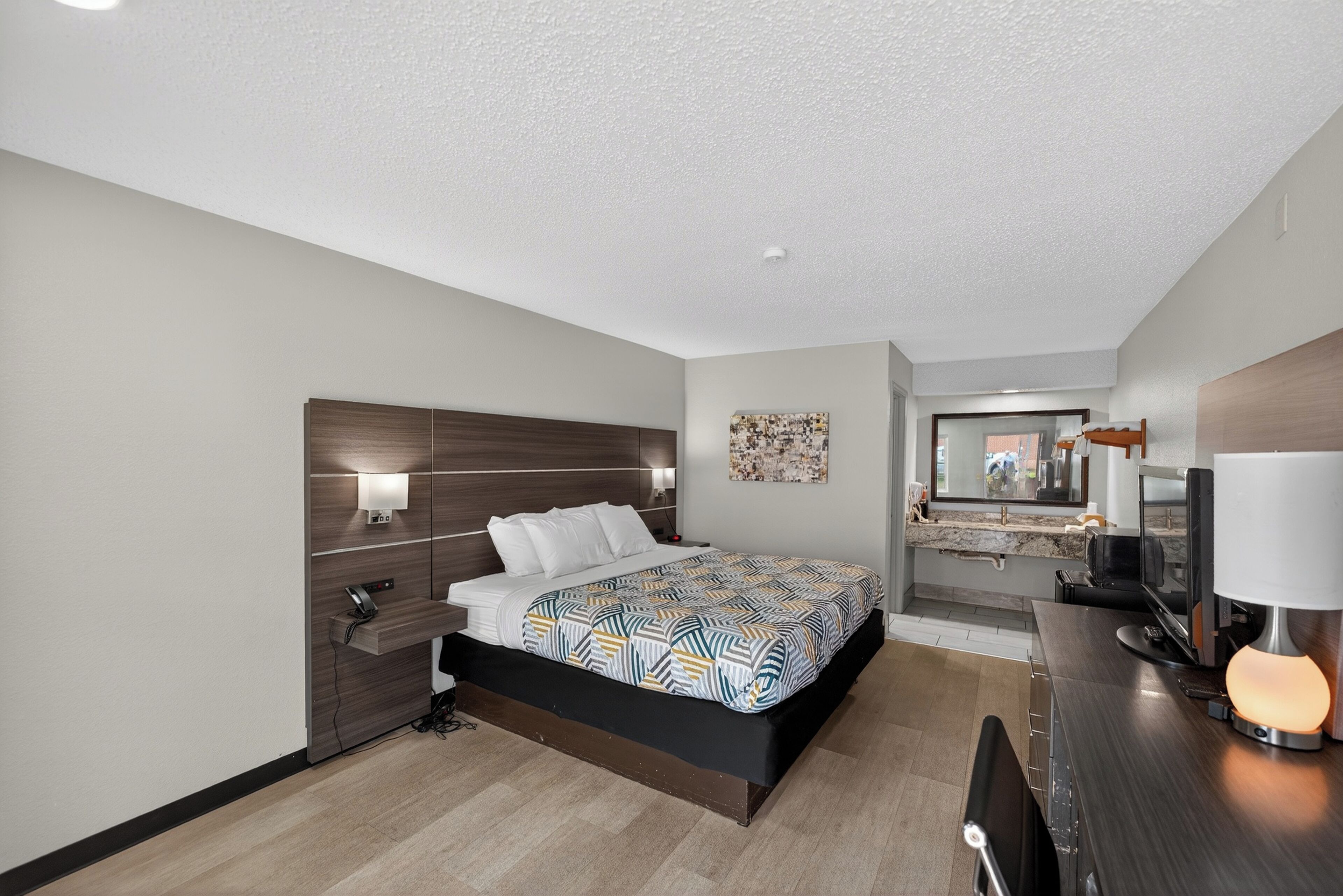 A modern hotel room with a bed, bedside table, lamp, and a small desk area with a computer.
