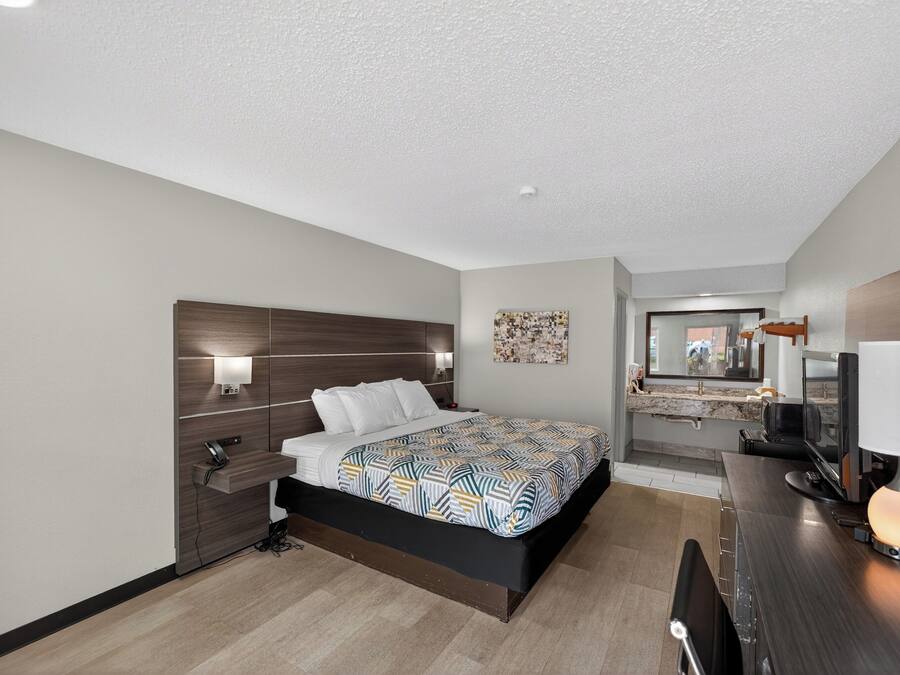 Ein modernes Hotelzimmer mit Bett, Nachttisch, Lampe und einer kleinen Arbeitsfläche mit Computer.
