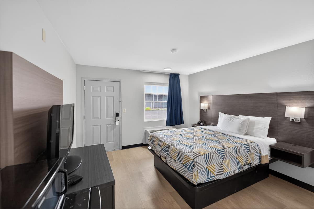 A hotel room with a bed, a desk, a TV, a window with curtains, and a wall-mounted lamp.