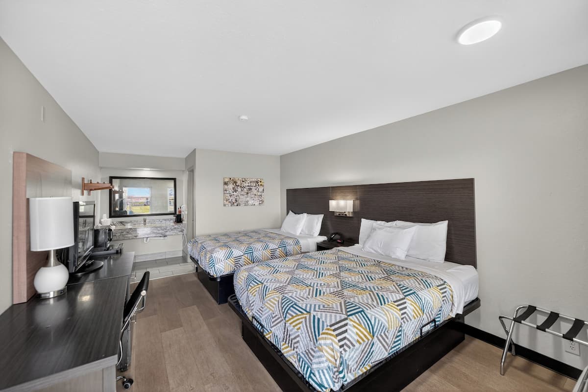 A hotel room with two beds, a desk with a lamp, a TV, and a bathroom visible in the background.