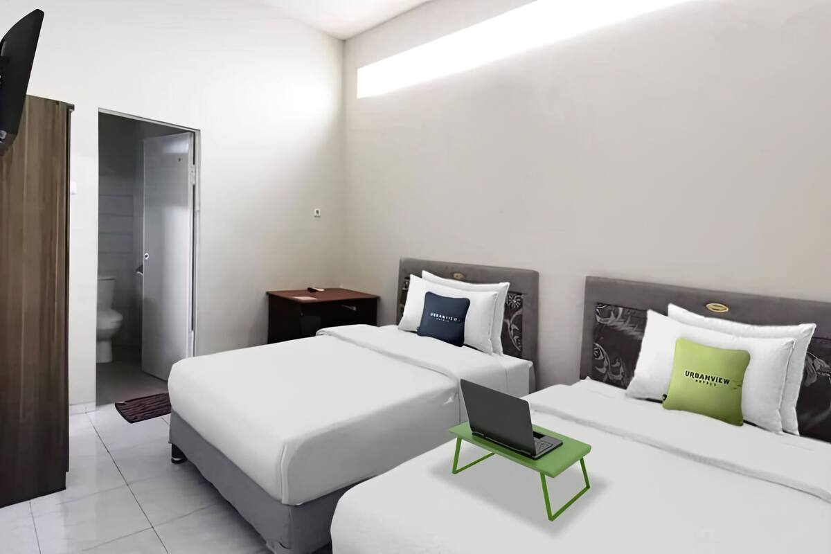 Une chambre d’hôtel avec deux lits, un ordinateur portable posé sur un support vert et une table de chevet avec une lampe.