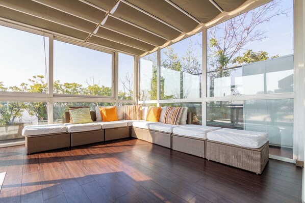 A covered outdoor seating area with a large window, wooden flooring, and a view of trees.