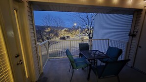 A covered balcony with a table and chairs, a view of a street and houses, and string lights.