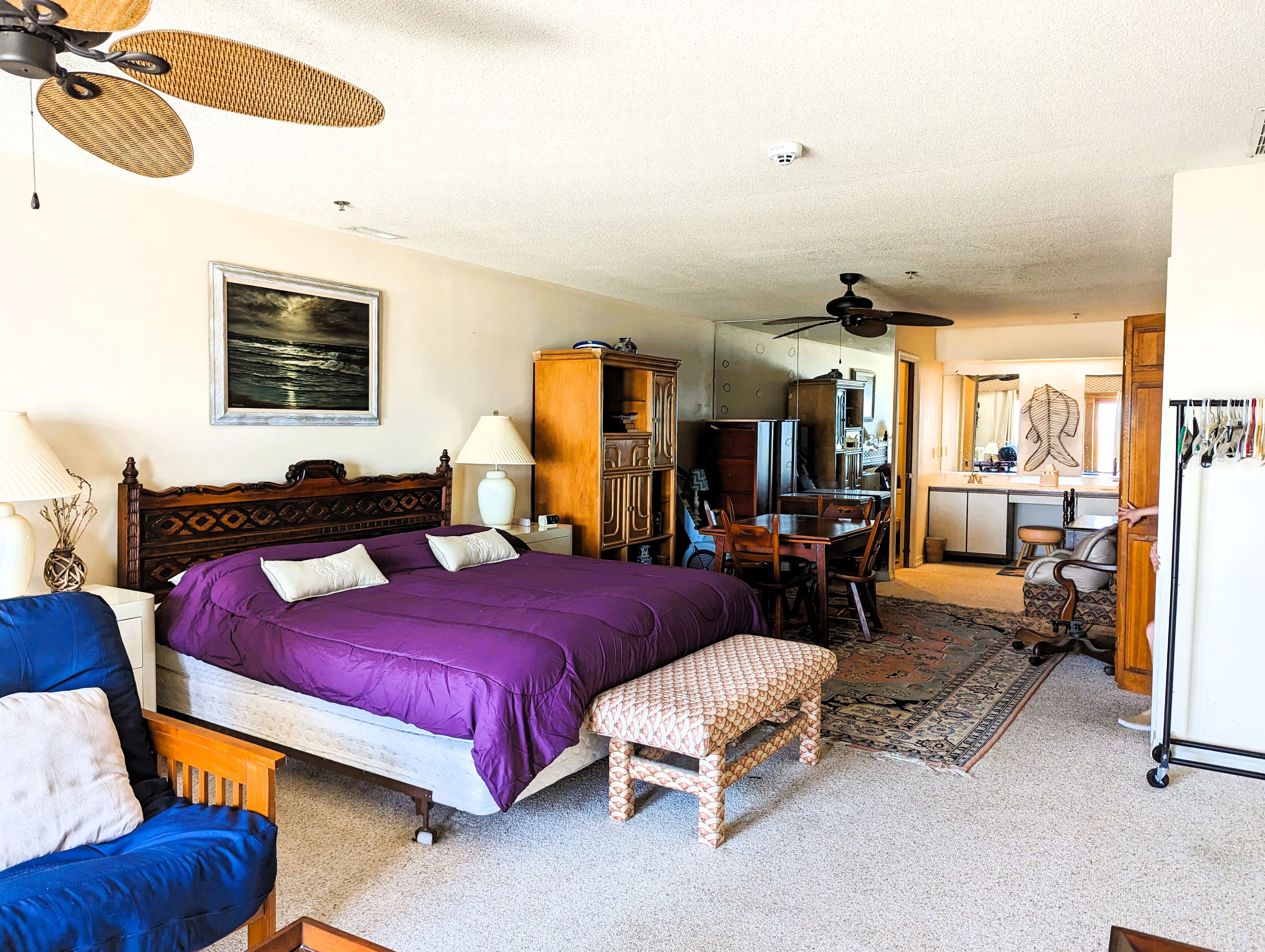 A bedroom with a large bed, a desk, and a chair. There is a ceiling fan, a lamp, and a painting on the wall.
