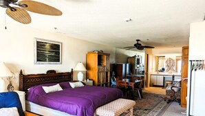 Une chambre avec un grand lit, un bureau et une chaise. On y trouve un ventilateur de plafond, une lampe et un tableau au mur.