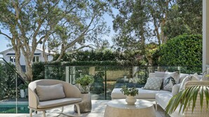 A modern outdoor seating area with a glass balustrade, a sofa, a chair, and a coffee table, surrounded by lush greenery and trees. - Uber Luxury 4 Bed Home with Pool in Mosman (Mosman)
