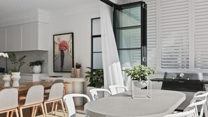 A modern kitchen with white cabinets, a dining area with a wooden table and white chairs, a large window with blinds, and a grill outside. - Uber Luxury 4 Bed Home with Pool in Mosman (Mosman)