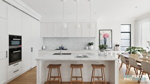 A modern kitchen with white cabinetry, a marble countertop, a built-in oven, and a dining area with wooden stools and a table. - Uber Luxury 4 Bed Home with Pool in Mosman (Mosman)