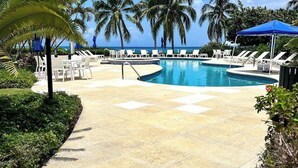 A pool area with lounge chairs and umbrellas, surrounded by palm trees and a clear sky. - Crescent Point Condo #9 - 3BR 7 Mile Beach Condo (George Town)