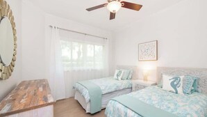 A bedroom with two beds, a ceiling fan, a large mirror, and a window with curtains. - Crescent Point Condo #9 - 3BR 7 Mile Beach Condo (George Town)