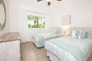 Ein Schlafzimmer mit zwei Betten, einem Schminktisch, einem Fenster mit Blick auf Grünflächen und einem Deckenventilator.