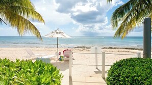 A beach with a white umbrella, lounge chairs, and a small kiosk. - Crescent Point Condo #9 - 3BR 7 Mile Beach Condo (George Town)