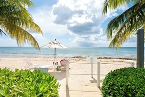 A beach with a white umbrella, lounge chairs, and a small kiosk. - Crescent Point Condo #9 - 3BR 7 Mile Beach Condo (George Town)