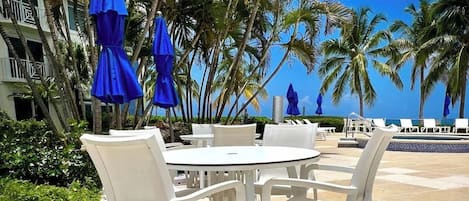 A poolside area with white lounge chairs and tables, blue umbrellas, and palm trees.