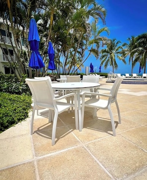 A poolside area with white lounge chairs and tables, blue umbrellas, and palm trees.