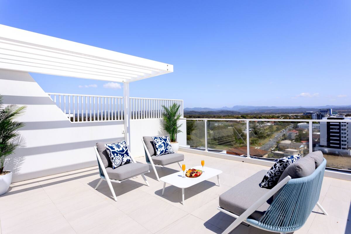 A rooftop terrace with white furniture, a white table with fruit and drinks, and a view of distant buildings and mountains.