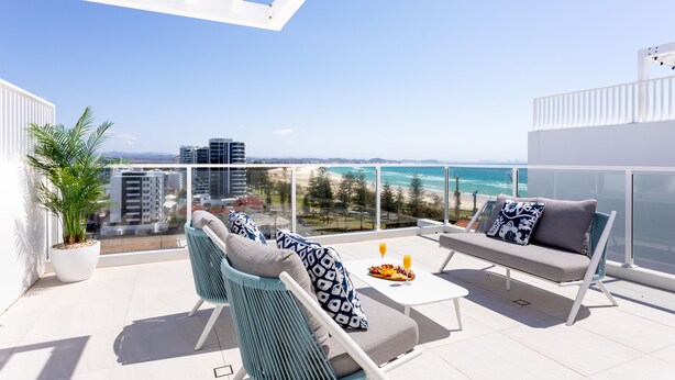 A balcony with a white railing, a potted plant, two chairs, a sofa, and a table with fruit and drinks. - The Beach Houses Holiday Villas (Coolangatta)