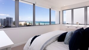 A modern bedroom with a large bed, white bedding, and a view of the ocean and buildings through large windows. - The Beach Houses Holiday Villas (Coolangatta)