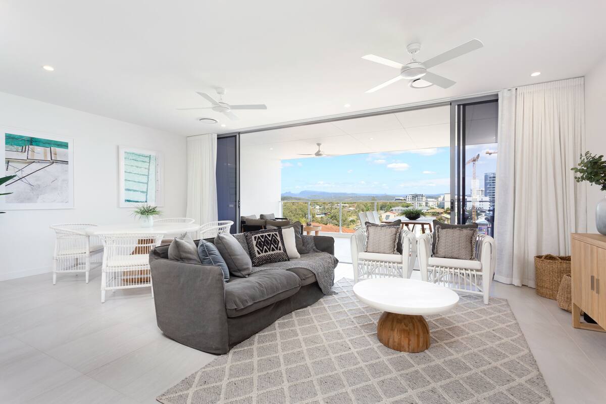 A modern living room with a grey sofa, white chairs, a round table, and large windows offering a city view.