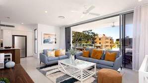 A modern living room with a sofa, ottoman, and coffee table, a kitchen area, and a large sliding glass door leading to an outdoor balcony. - The Beach Houses Holiday Villas (Coolangatta)