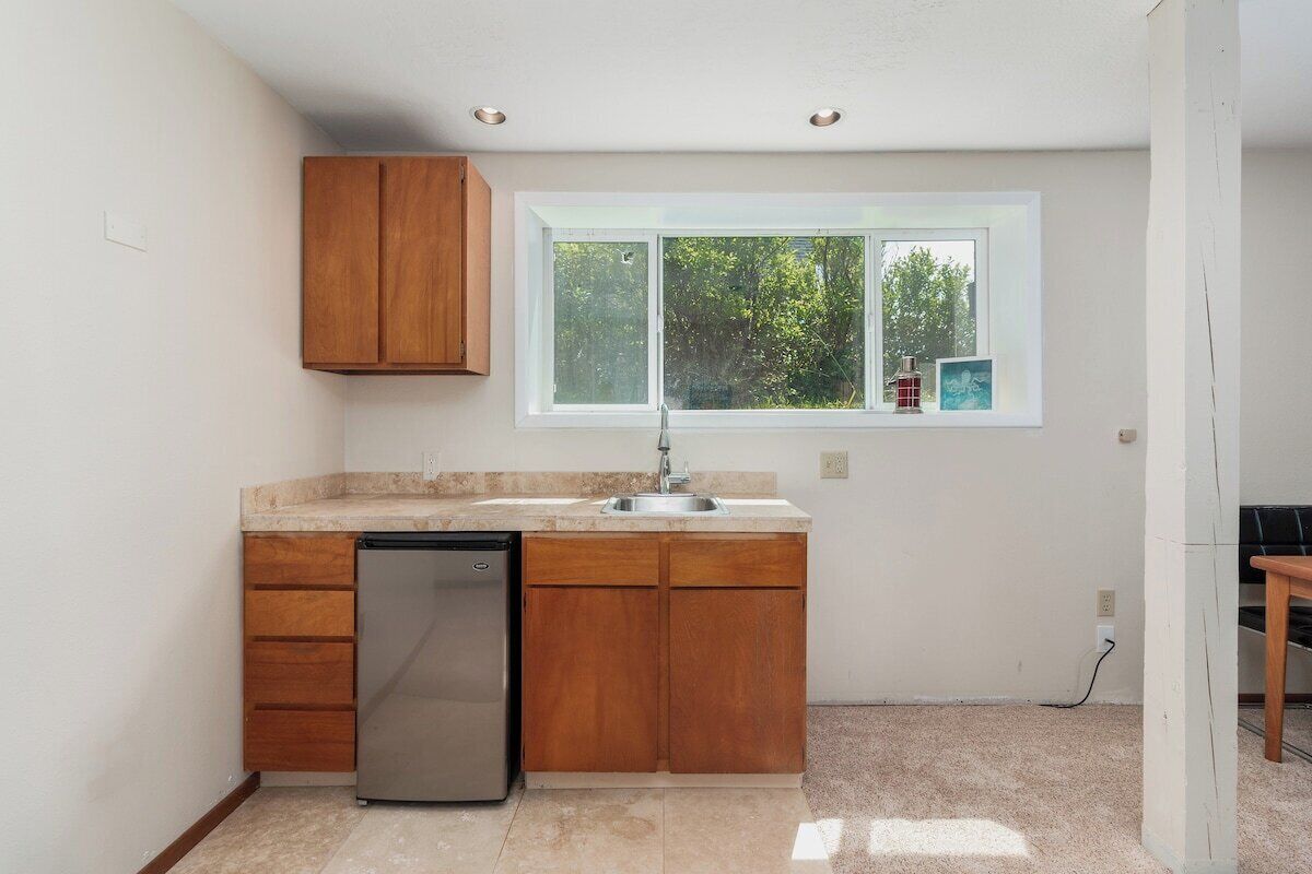 A compact kitchen with wooden cabinets, a stainless steel dishwasher, and a window with a view of greenery.