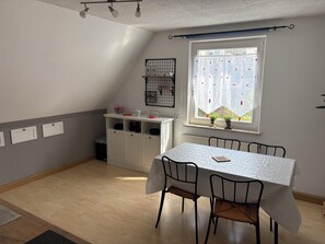 Un coin repas comprenant une table, des chaises, une fenêtre avec des rideaux, un radiateur et un buffet blanc contenant de la vaisselle.