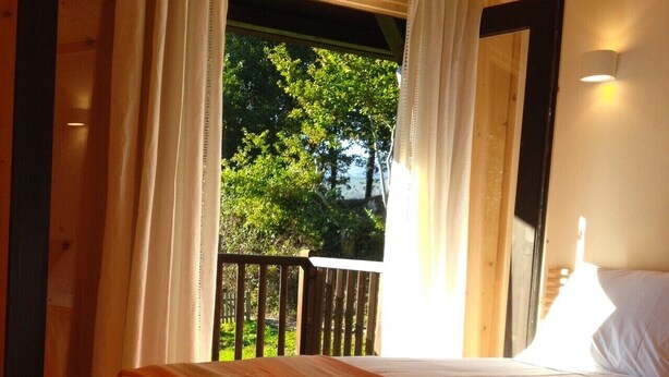 A bedroom with a bed, a window with curtains, a view of greenery, and a wooden deck outside. - Treehouse Quinta Lamosa (Arcos de Valdevez)