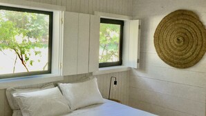 Een slaapkamer met een bed, twee ramen, een geweven wanddecoratie en een zichtbare stopcontact.