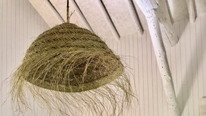 Een kamer met een witte bank, een hangende geweven lamp, een potplant en een klein houten krukje.