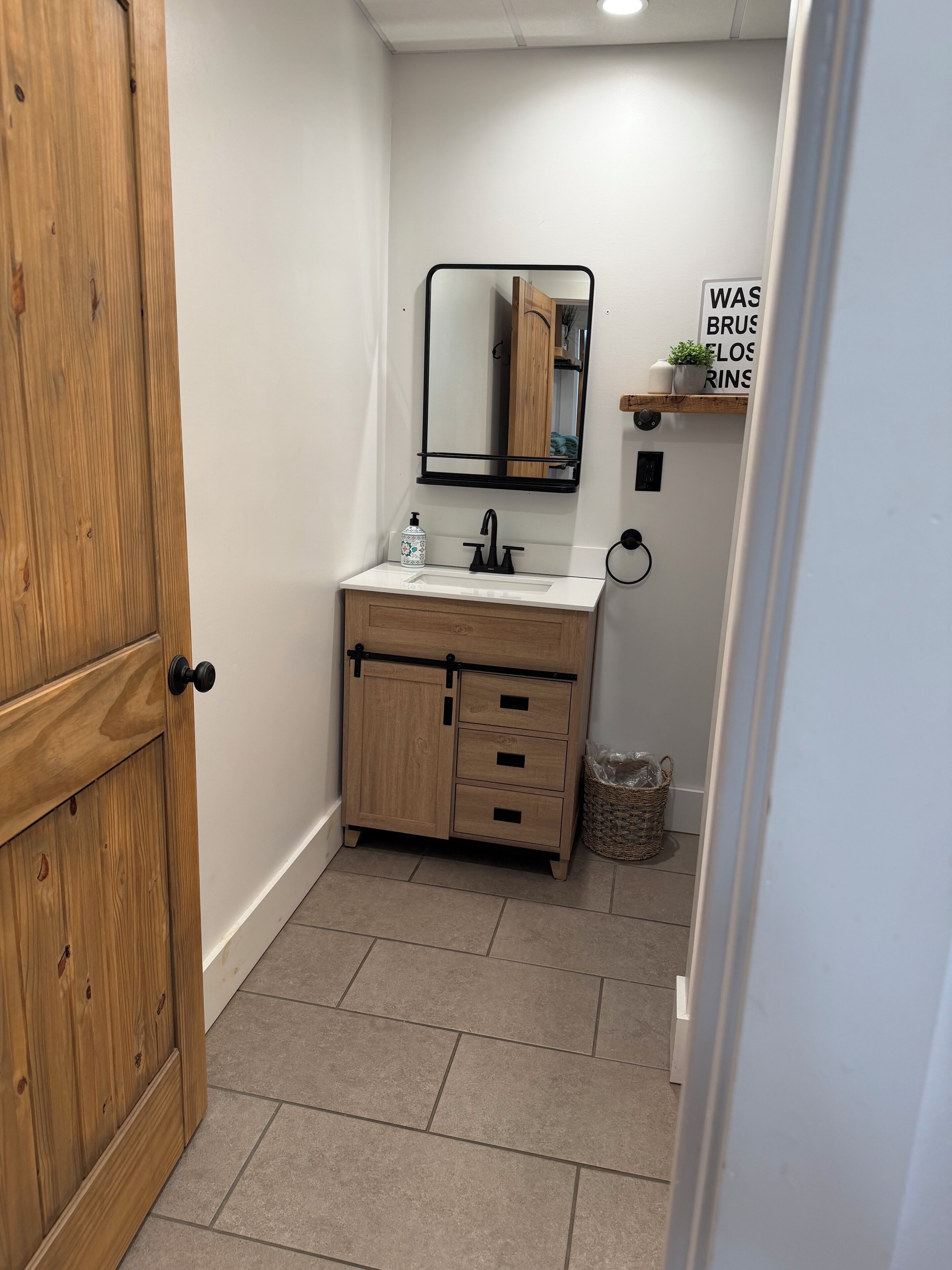 Ein Badezimmer mit Holzschrank, weißem Waschbecken, schwarzem Wasserhahn, einem Spiegel und einem Regal mit einer Pflanze.