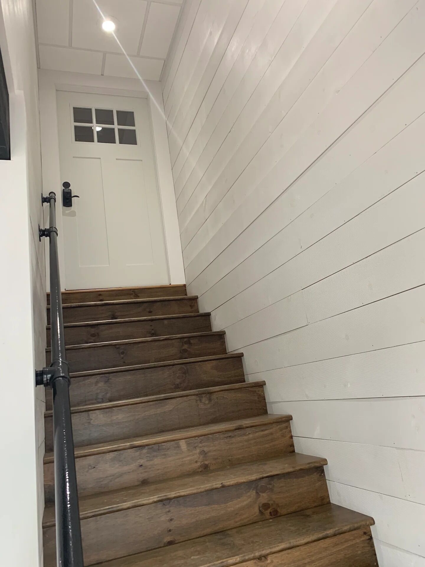 Una escalera con escalones de madera y barandilla negra, que lleva a una puerta blanca con ventana.
